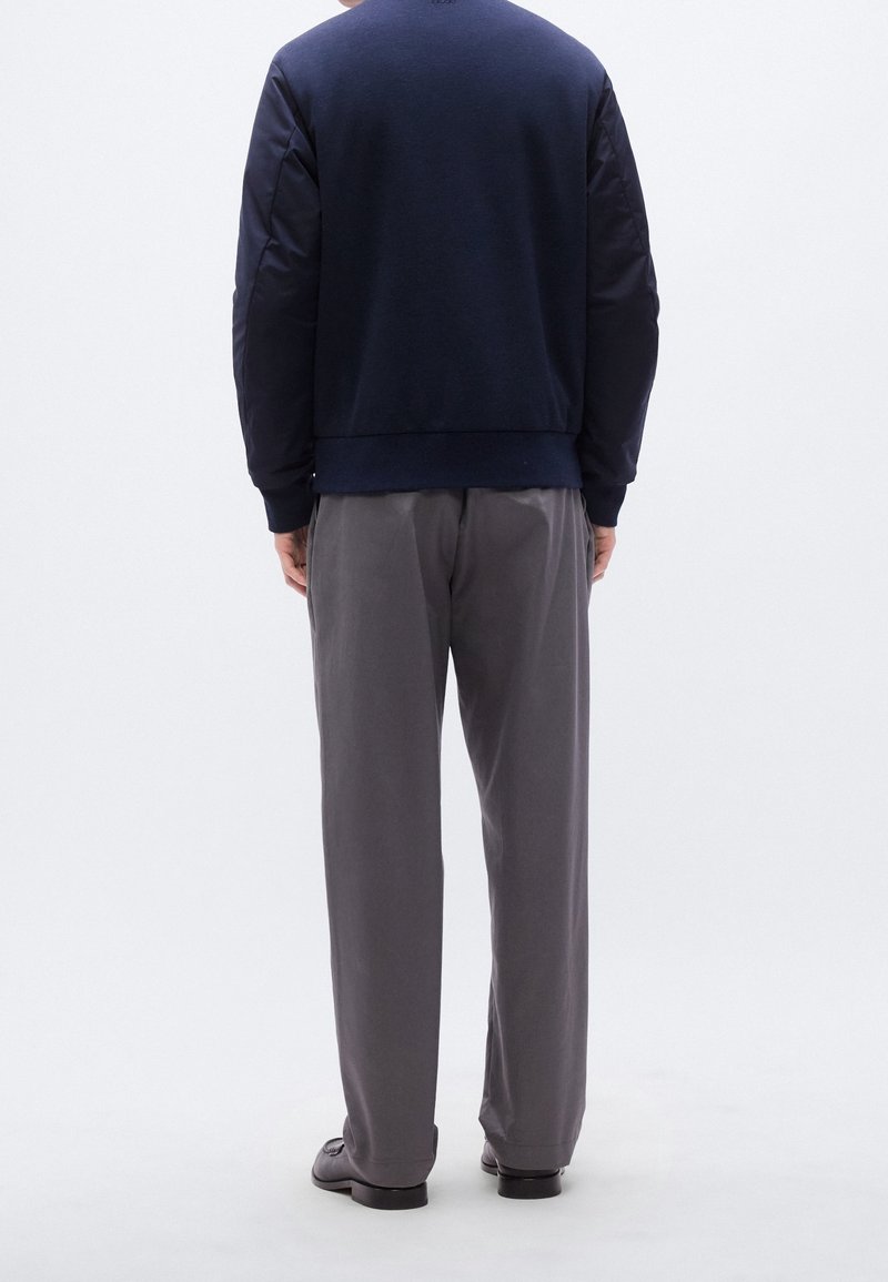 Personne debout de dos portant une veste bleu foncé, un pantalon gris et des chaussures noires sur un fond blanc uni.