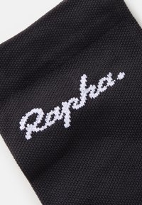 Crne biciklističke čarape s reljefnim pletenim uzorkom i bijelim izvezenim logom "Rapha." blizu vrha.