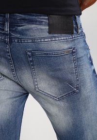 Dżinsy typu denim z gradientowym niebieskim wyblaknięciem, pięciopocketowym designem oraz czarną skórzaną naszywką w pasie, z subtelnymi detalami w szyciu.