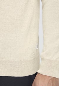 Suéter de punto beige claro con escote redondeado, puños y dobladillo acanalados. Pequeña etiqueta de la marca en la costura lateral. Textura suave y ajuste casual.