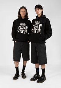 Des sweats à capuche noirs avec un design graphique blanc, associés à des shorts en denim foncé. Les deux modèles portent des chaussures et des chaussettes noires, se tenant ensemble devant un fond neutre.
