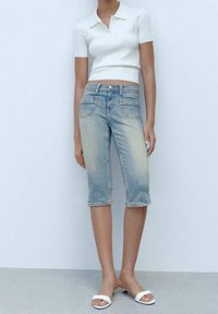 Pantalones capri de mezclilla azul claro con acabado desgastado, cinco bolsillos y cierre de cremallera, combinados con una camiseta polo blanca ajustada de tejido acanalado.