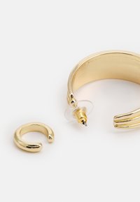 Brincos de metal dourado com um design de argola espessa e um pequeno pino em forma de C. A argola apresenta um acabamento texturizado e um suporte transparente.