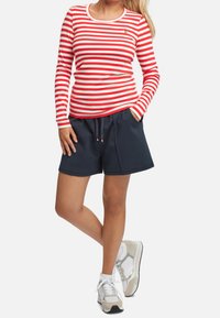 Rot-weißer gestreifter Langarm-Top kombiniert mit marineblauen Shorts mit elastischem Bund, seitlichen Taschen und hellgrauen Sportschuhen.