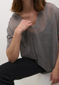 Top grigio lucido a scollo a V con maniche corte, caratterizzato da una texture trasparente e una vestibilità ampia, abbinato a pantaloni neri.
