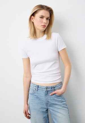 Junge Frau mit schulterlangem blondem Haar, die ein eng anliegendes weißes Kurzarmshirt und hellblaue Jeans vor einem einfarbigen weißen Hintergrund trägt.