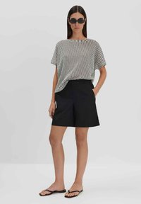 Gestreepte zwart-witte, loszittende top, gecombineerd met hoog opgesneden zwarte shorts en minimalistische zwarte slippers, tegen een neutrale achtergrond.