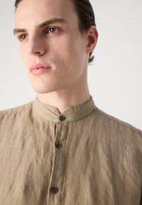 Camisa de botones hecha de una tela texturada de color marrón claro con cuello bandana y botones oscuros, que presenta un ajuste relajado y sutiles arrugas.