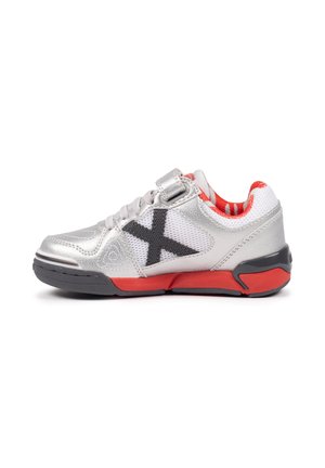 Zapatilla plateada y blanca con una "X" negra en el lateral, suela roja y negra, y interior a rayas rojas, vista desde el lado exterior.
