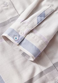 Poignet de chemise à manches longues présentant un motif à carreaux bleu clair et gris avec des boutons blancs et une couture détaillée sur un tissu texturé.