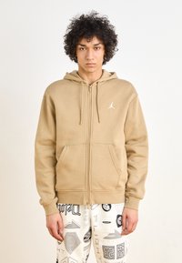 Sudadera con cremallera color beige hecha de una tela suave, con un bolsillo tipo canguro en la parte frontal y un logo blanco. Combinada con pantalones blancos estampados.