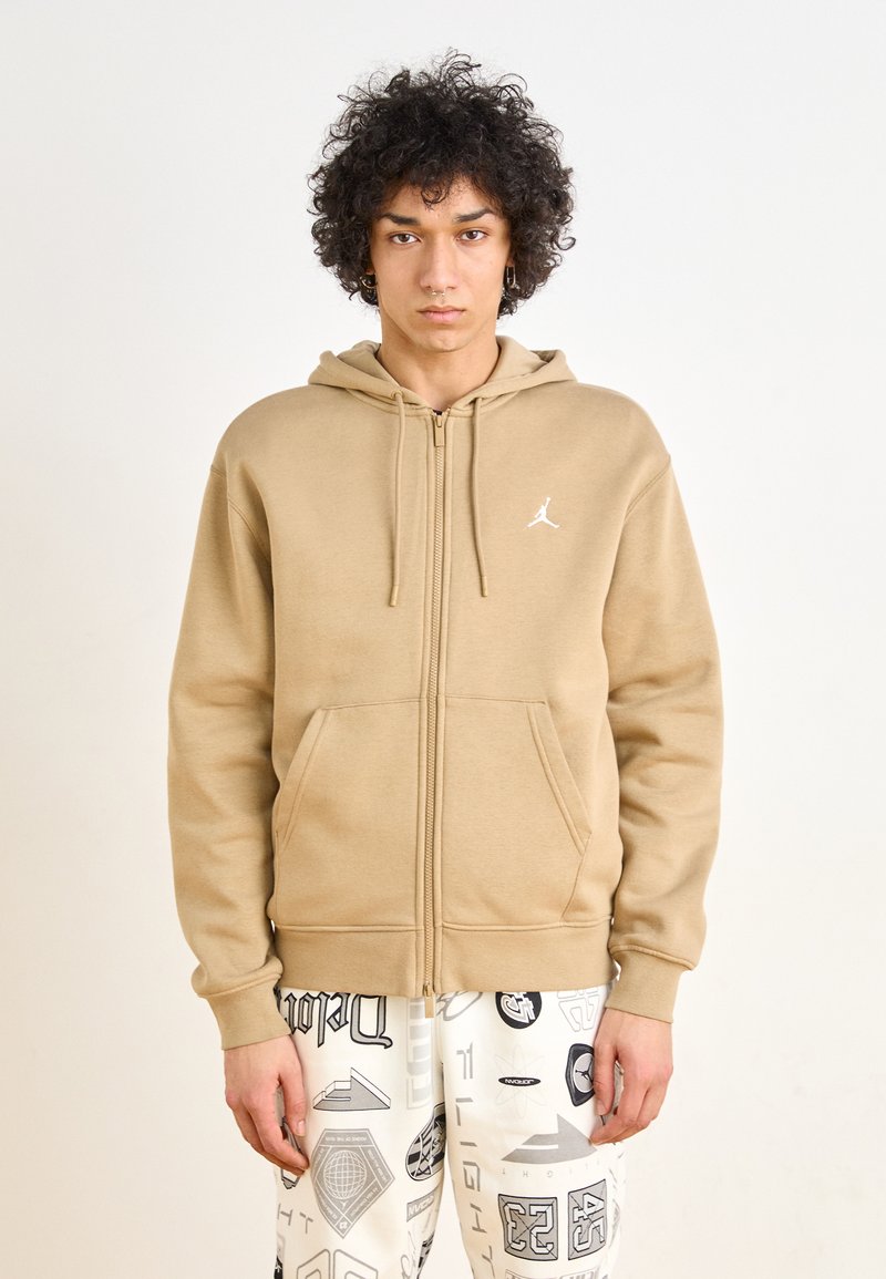 Sudadera con cremallera color beige hecha de una tela suave, con un bolsillo tipo canguro en la parte frontal y un logo blanco. Combinada con pantalones blancos estampados.