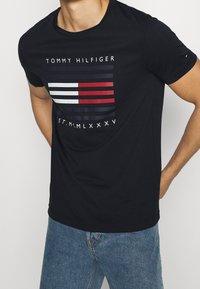 Camiseta de algodón azul marino con un gráfico de franjas horizontales en rojo, blanco y azul oscuro, con el texto "TOMMY HILFIGER" arriba.