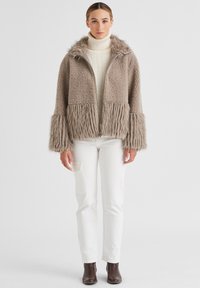 Faux-Fur-Reißverschlussjacke in hellbeige mit strukturiertem Stoff und flauschigem Saum und Bündchen, kombiniert mit weißen Hosen und dunkelbraunen Stiefeletten.