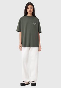 Oliivinvihreä ylisuuri puuvillainen t-paita, jossa on lyhyet hihat, pyöreä pääntie ja valkoinen logotunnus. Yhdistetty valkoisiin leveälahkeisiin housuihin ja kenkiin.