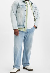 Giacca di jeans blu chiaro, t-shirt bianca, camicia a righe verde e gialla sotto, jeans blu chiaro e scarpe bianche. Outfit casual.