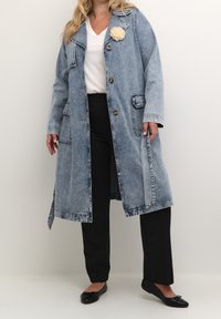 Žena ve světle modrém džínsovém dlouhém kabátu s textilní květinovou broží, bílém tričku, černých kalhotách a černých balerínách na jednoduchém pozadí.