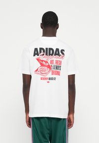 Hvid bomulds t-shirt med et grafisk design på ryggen, der har fed tekst i sort og rødt, som viser en pizzaboks og leveringsdetaljer.