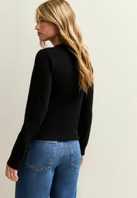 Schwarzer, gestrickter Pullover mit langen Ärmeln, hohem Kragen und tailliertem Schnitt, kombiniert mit blauen, hochtaillierten Jeans und einer glatten Textur.