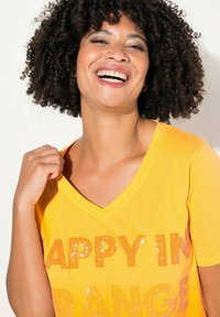 T-shirt en coton jaune avec un col V, portant l'inscription "HAPPY IN ORANGE" en texte de sequins orange. Texture douce et coupe décontractée.