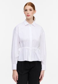 Camicia bianca con bottoni, vita arricciata, maniche lunghe e polsini. Tessuto leggero e morbido con colletto classico.