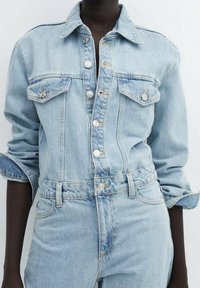 Combinaison en denim bleu clair avec un col à revers, des manches longues retroussées, deux poches poitrine, une fermeture boutonnée devant et une ceinture à la taille.