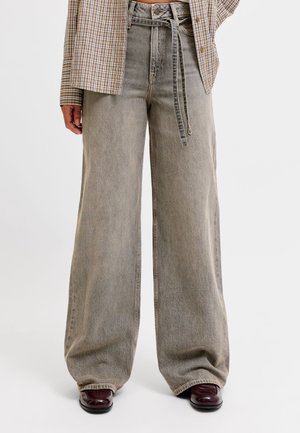Personne portant un jean gris taille haute à jambes larges avec une ceinture en tissu, une chemise à carreaux et des chaussures foncées et brillantes sur fond blanc.