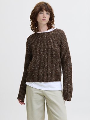 Jong persoon met schouderlang roodachtig haar, een dikke bruine gebreide trui over een wit overhemd en beige broek, staand tegen een witte achtergrond.