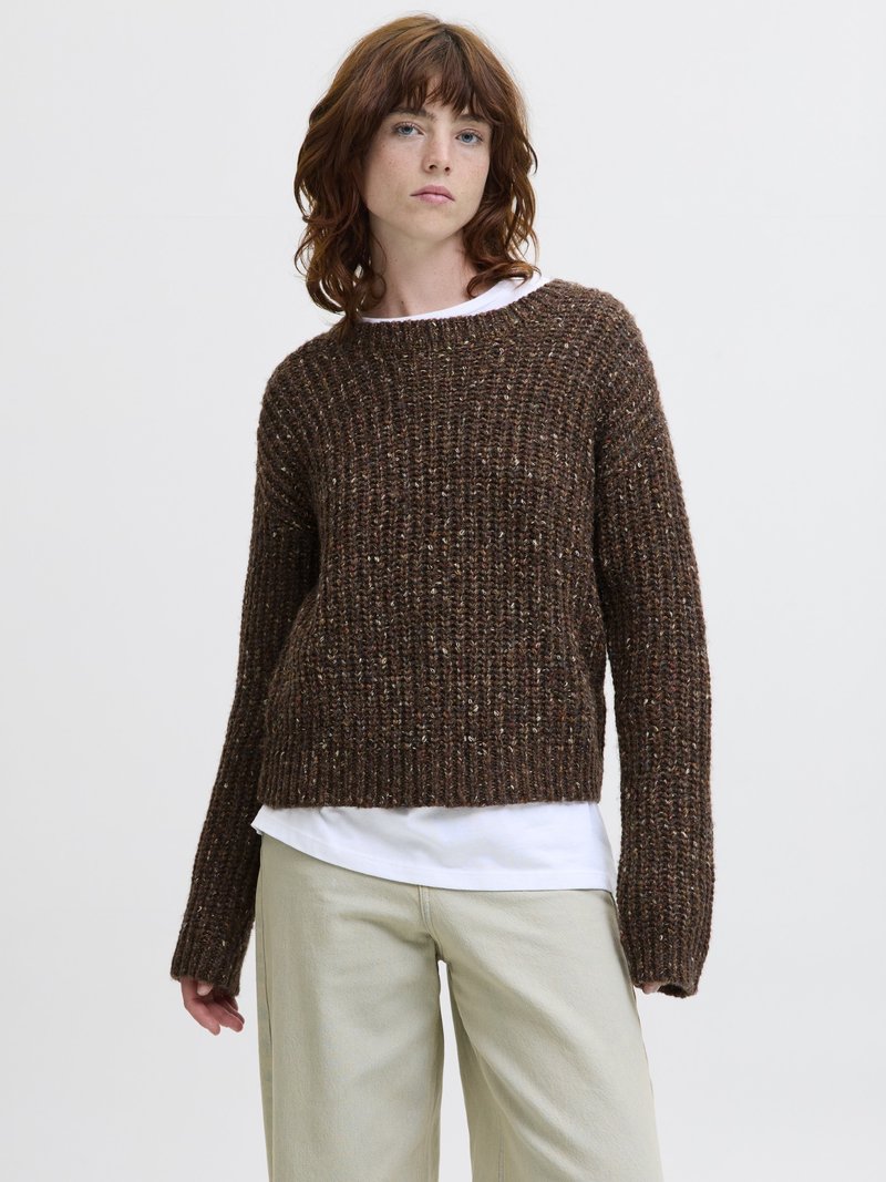 Jeune personne aux cheveux roux mi-longs portant un gros pull en laine marron sur une chemise blanche et un pantalon beige, debout devant un fond blanc.