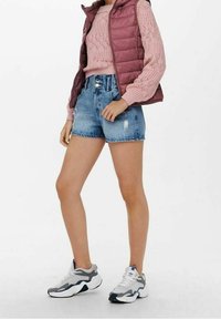 Pikowana kamizelka w głębokim bordo na jasnoróżowym dzianinowym swetrze, zestawiona z przetartymi szortami jeansowymi i szarymi butami sportowymi z białymi podeszwami.