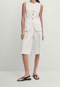Witte gestreepte mouwloze vest met knopen, gecombineerd met knielange witte shorts. Zwarte sandalen maken de outfit compleet en tonen een strak ontwerp.
