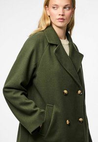 Dubbelzijdige olijfgroene jas van textuurstof met een omgeslagen kraag, gouden knopen en zijzakken.