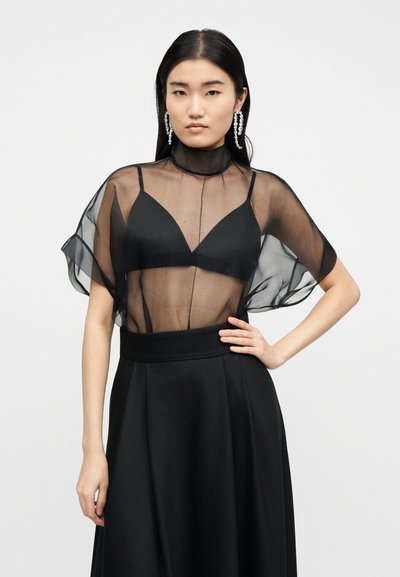 Γυναίκα ντυμένη με μαύρη διαφανή μπλούζα πάνω από μαύρο σουτιέν, μακριά μαύρη φούστα και κρεμαστά σκουλαρίκια με κρύσταλλα, στέκεται με το ένα χέρι στη μέση.