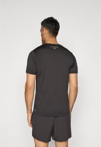 T-shirt de sport noir à manches courtes avec un col rond, présentant un logo blanc dans le haut du dos. Tissu lisse et design ajusté.