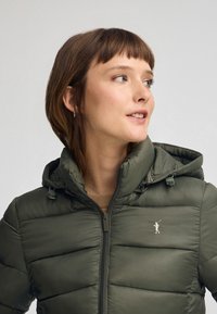 Olivgrön puffjacka med huva, med kviltat mönster, framdragkedja och en liten guldfärgad logotyp på bröstet.