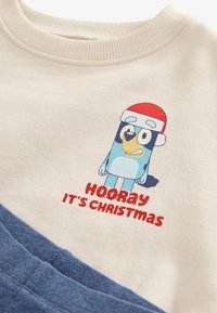 Sweter w kolorze ecru z rysunkowym ptakiem w czerwonej czapce Mikołaja oraz napisem "HOORAY IT'S CHRISTMAS" na czerwono, z niebieskimi akcentami i prążkowanym dekoltem.
