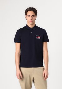 Mørkeblå polo-skjorte med krage, korte ermer og en liten brodert logo. Båret med lysebeige bukser. Enkel og tettsittende design.