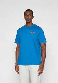 Mėlynas medvilninis marškinėlis su apvaliu iškirpimu ir trumpomis rankovėmis, kuriame kairėje krūtinės pusėje yra mažas daugiaspalvis siuvinėjimas.