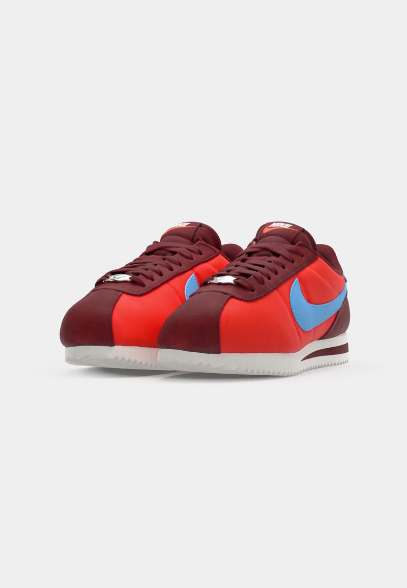 Zapatillas rojas y burdeos con un swoosh azul de Nike, parte superior texturizada y suela de goma blanca. Presentan cordones y una etiqueta de marca en la lengüeta.
