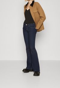 Brun kappa med svart krage, svart topp och mörkblå utsvängda jeans. Svarta klumpiga klackstövlar. Minimalistisk design, figurnära silhuett.