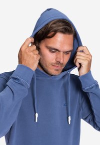 Blauer Kapuzenpullover aus weichem Stoff, mit Kordeln, gerippten Bündchen und einem kleinen Logo auf der Brust. Texturierte Kapuzen-Details.
