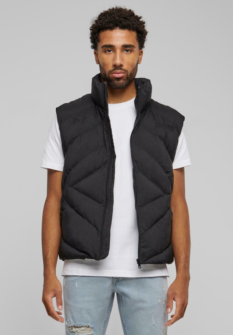 Schwarze Steppweste mit Stehkragen, gestepptem Chevron-Muster und Frontreißverschluss. Getragen über einem weißen T-Shirt, kombiniert mit hellblauen Jeans.