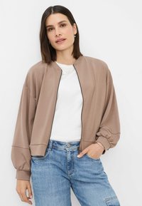 Brown bomber jacket with ribbed cuffs, zip front, and relaxed fit, worn over a white t-shirt and accompanied by light blue jeans.