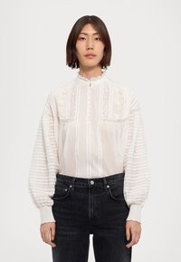 Hvid bluse med broderi, knaplukning foran, høj hals og voluminøse ærmer; parret med mørke jeans; gennemsigtig og struktureret stof.