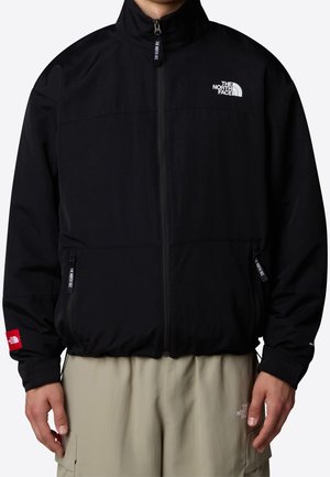 Veste noire zippée North Face portée sur un pantalon beige, avec des logos visibles de la marque sur la poitrine, les manches et les curseurs de fermeture éclair.