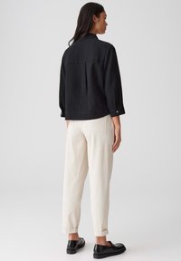 Zwarte gemêleerde shirt met een relaxte pasvorm en knoopmanchetten, gecombineerd met witte broeken en zwarte schoenen. Achteraanzicht toont geen belangrijke details.