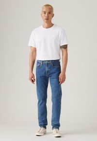 Hombre con cabello rubio corto que lleva una camiseta blanca, jeans azules, zapatillas blancas y un tatuaje en la parte superior del brazo izquierdo, de pie contra un fondo liso.