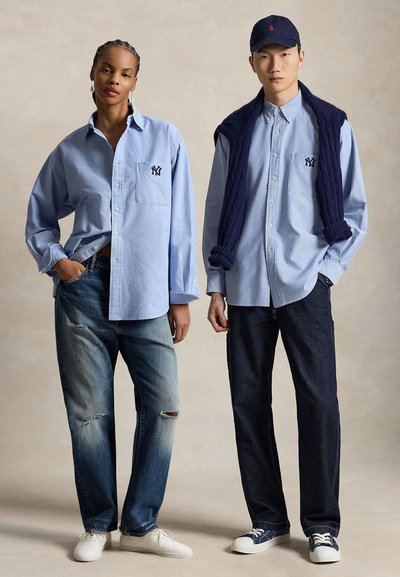 Zwei Models tragen hellblaue Hemden mit Knöpfen und „NY“-Logos; eines kombiniert das Hemd mit zerrissenen Jeans und weißen Schuhen, das andere mit dunklen Jeans, Sneakers und einem dunkelblauen Pullover.