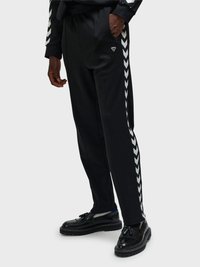 Person trägt schwarze Jogginghose mit weißen Chevron-Streifen an den Seiten und schwarze Loafer mit Quasten, steht vor einem schlichten Hintergrund.