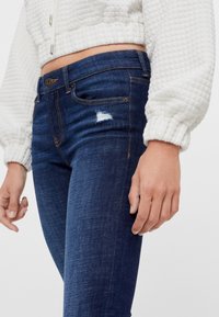 Mörkblå denimjeans med slim fit, med en liten trasig detalj, tillsammans med en vit texturerad kort jacka.